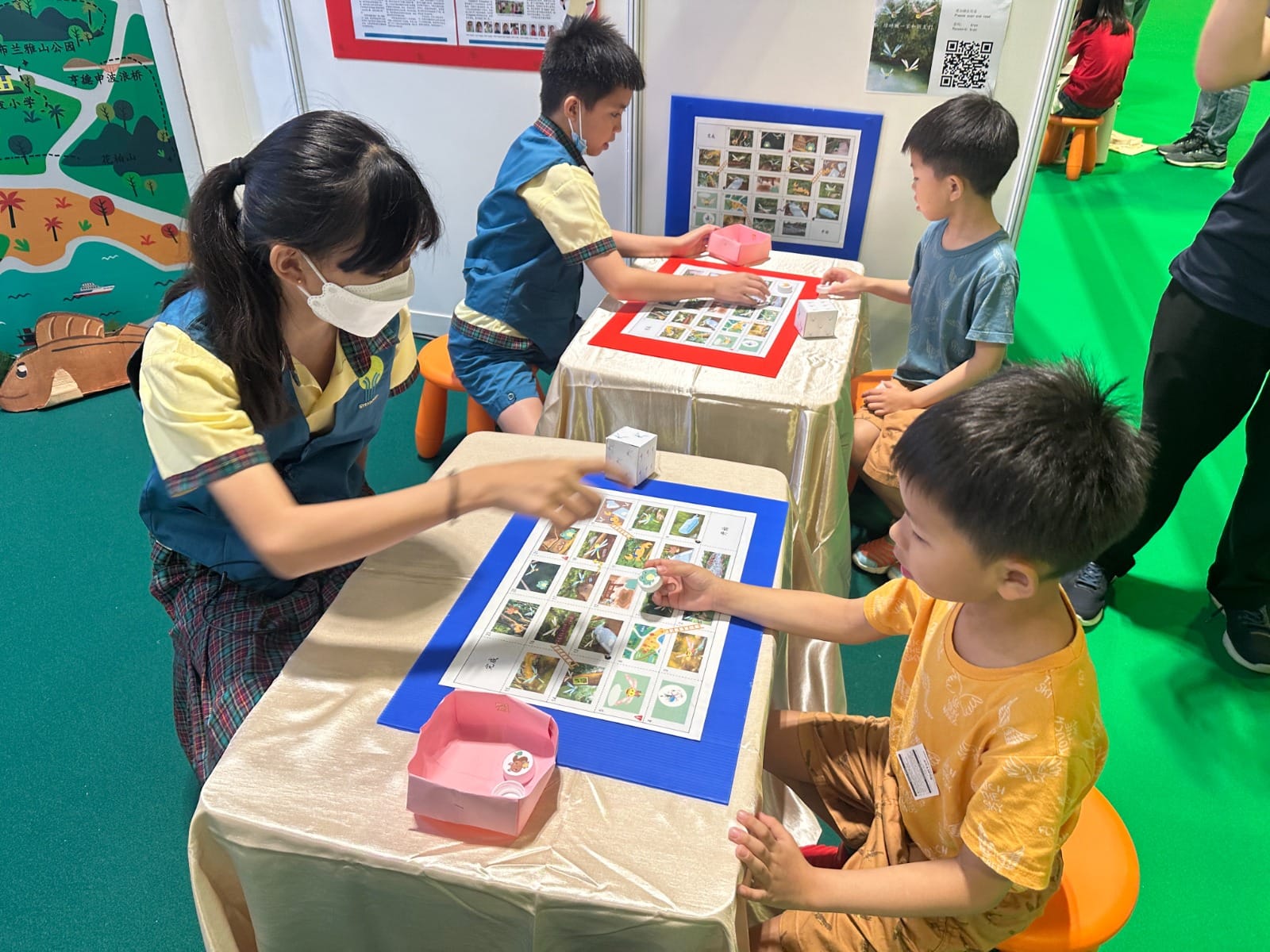 A masked woman and young children play a nature-themed board game at tables in a colorful indoor exhibit space.